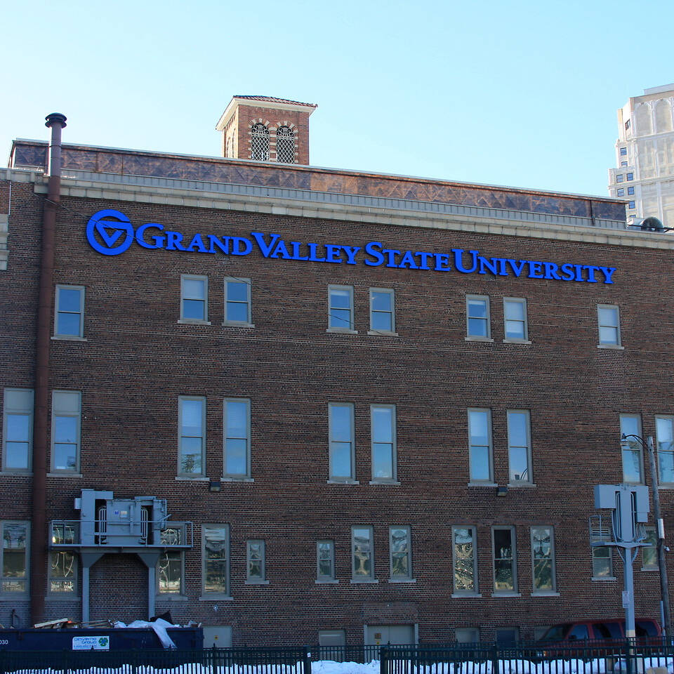 A brick building with "Grand Valley State University" signage stands in a snowy urban setting, with tall modern skyscrapers visible in the background.